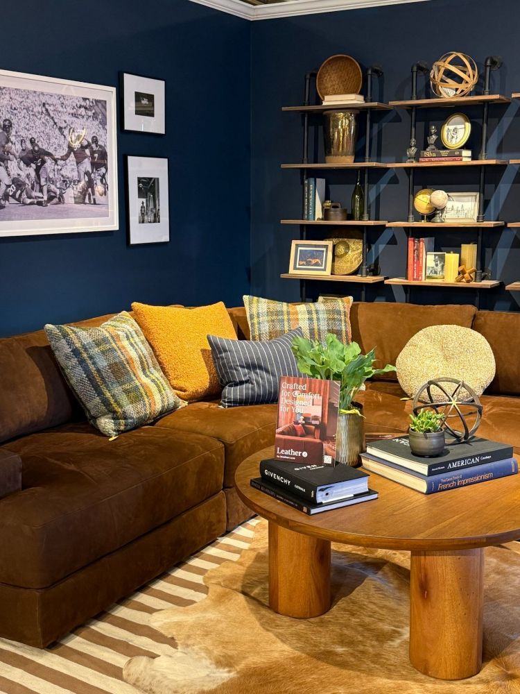 Stunning Rust Colored Sofa with wool textured throw pillows in front of a deep dark blue wall with a brown wooden coffee table with magazines and books, with table plants and a golden sphere. 